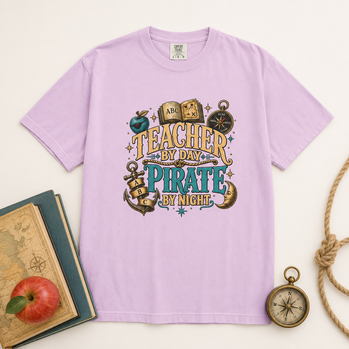 Teacher by Day Pirate by Night Comfort Colors tee laid flat in a light orchid color beside books and a compass