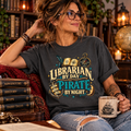 Woman in glasses relaxing in a cozy library, wearing a charcoal Librarian by Day Pirate by Night t-shirt with gold and teal lettering