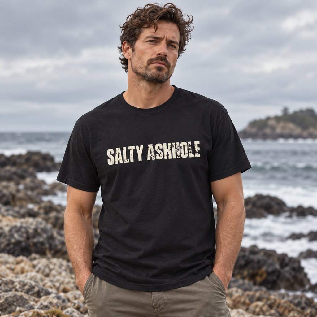 Man on a rocky shoreline wearing a black Salty Ashhole tee with distressed cream lettering across the chest