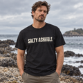 Man on a rocky shoreline wearing a black Salty Ashhole tee with distressed cream lettering across the chest