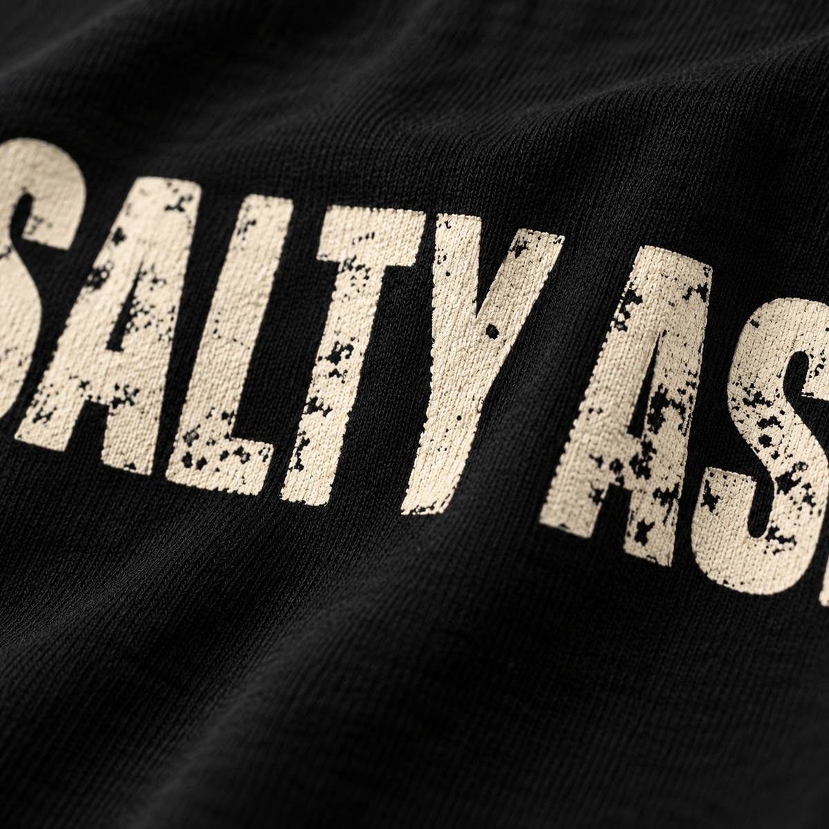 Close-up of the worn, distressed SALTY ASHHOLE lettering printed on a black tee