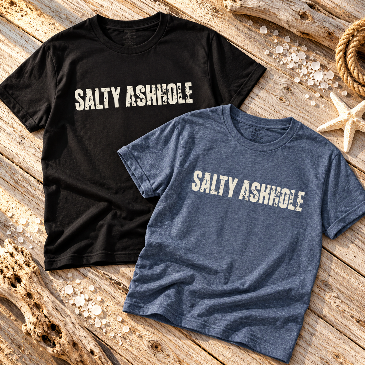 Black and heather blue Salty Ashhole tees laid on driftwood with rope and a starfish