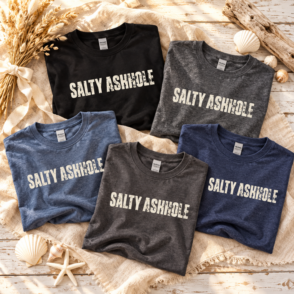 Five folded Salty Ashhole tees in all five colors on a beach blanket with seashells