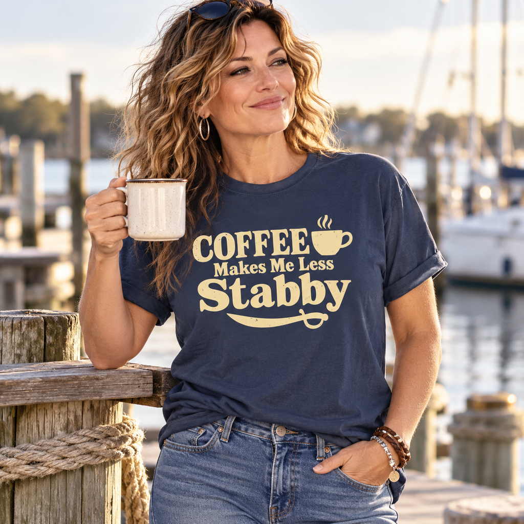 Woman holding a coffee mug on a marina dock, wearing a navy Coffee Makes Me Less Stabby Comfort Colors t-shirt