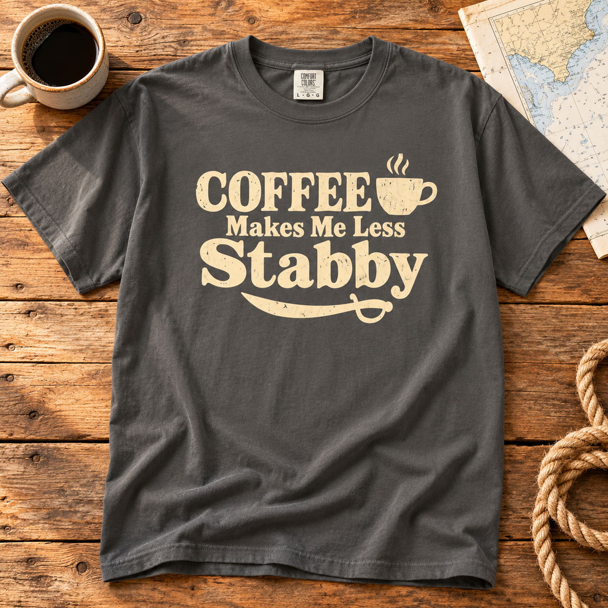 Pepper Coffee Makes Me Less Stabby t-shirt laid flat on a wooden table with a cup of coffee, a map and rope