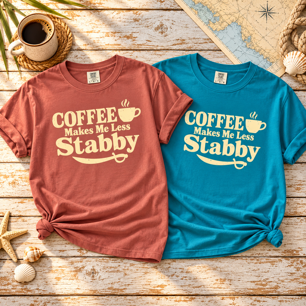 Two Coffee Makes Me Less Stabby t-shirts in Cumin and Topaz Blue laid flat on weathered wood with coffee and seashells