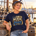 Woman in a navy Nurse by Day Pirate by Night t-shirt with PIRATE in ceil blue, standing on a dock by a tall ship at sunset