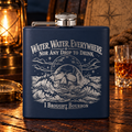 Navy hip flask engraved with Water, Water, Everywhere, Nor Any Drop to Drink, a skull rising from stormy waves and I Brought Bourbon at the bottom, on a wooden table by lantern light with a glass of bourbon