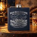 Navy hip flask engraved with Water, Water, Everywhere, Nor Any Drop to Drink, a skull rising from stormy waves and I Brought Whiskey at the bottom, on an old wooden table with a lantern, a compass and a glass of whiskey