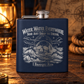 Navy hip flask engraved with Water, Water, Everywhere, Nor Any Drop to Drink, a skull rising from stormy waves and I Brought Rum at the bottom, on a wooden table with a lantern, rope and a glass of rum